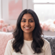 Smiling young South Asian woman with dark hair, wearing a light top, against a soft pink and white background.